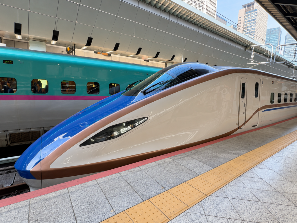 Blue-and-copper Shinkansen beside a green Shinkansen at a station platform